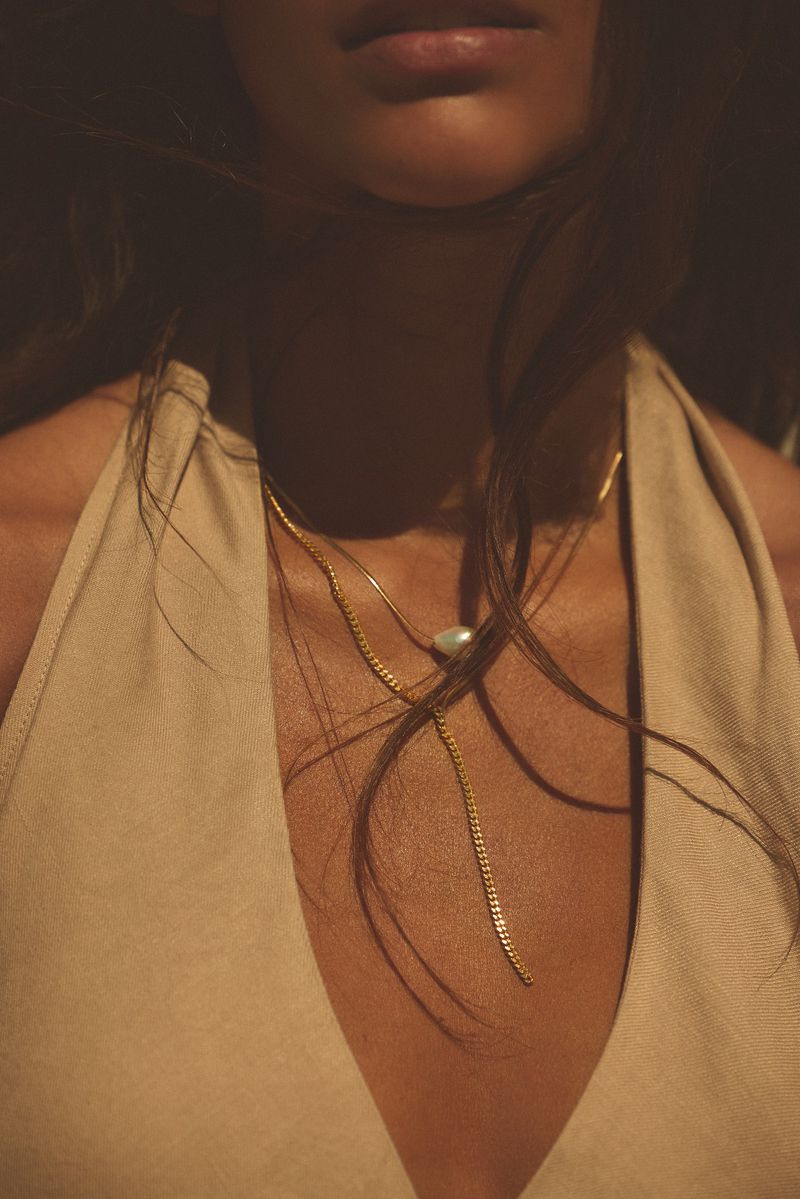 Close-up of a woman wearing a low-cut beige halter top and gold necklaces in soft natural lighting, focusing on her chest and neckline.