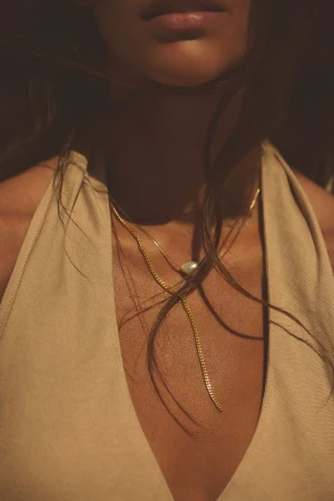 Close-up of a woman wearing a low-cut beige halter top and gold necklaces in soft natural lighting, focusing on her chest and neckline.