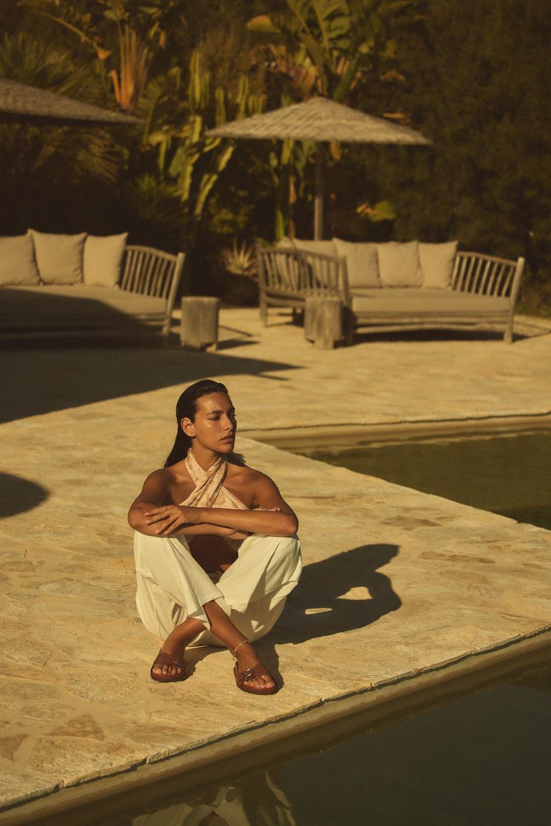 Adult female sitting cross-legged outdoors on a stone patio near a swimming pool and tropical plants in a fashion lifestyle editorial.
