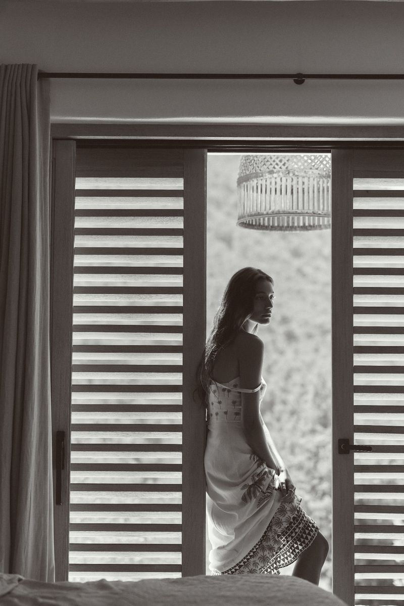 Monochrome photo of a woman in a dress standing in an open doorway, leaning against the frame, with a natural, blurred background.
