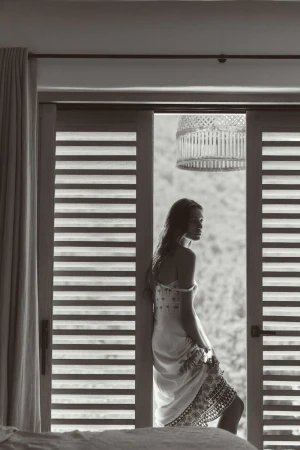 Monochrome photo of a woman in a dress standing in an open doorway, leaning against the frame, with a natural, blurred background.