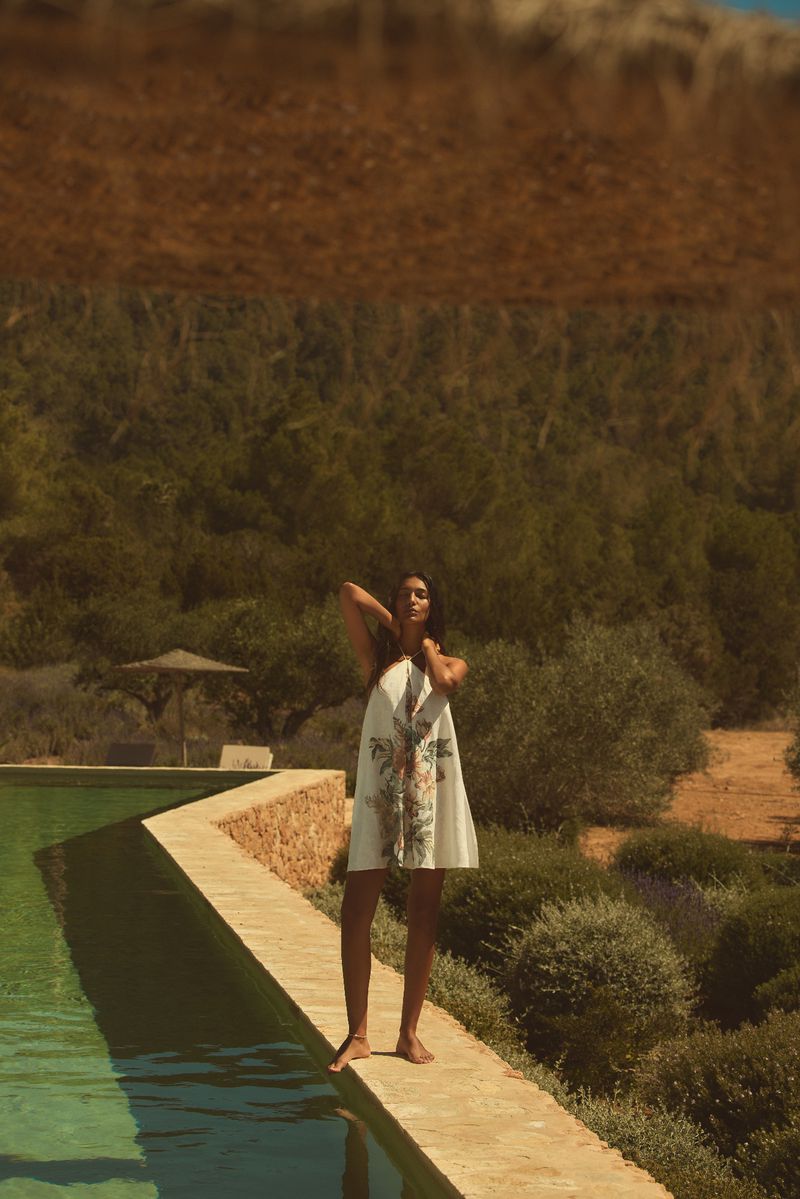 Woman poses in white floral dress on stone path near pool in sunny outdoor setting, lush greenery and hillside backdrop.