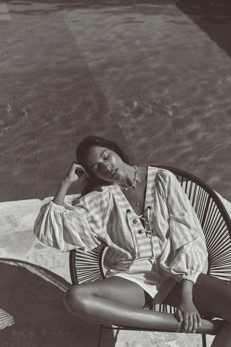 Monochrome photo of a woman in a striped shirt seated by a pool, eyes closed, bathed in sunlight, creating a relaxed and serene scene.