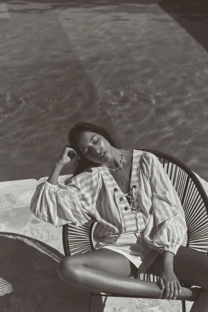 Monochrome photo of a woman in a striped shirt seated by a pool, eyes closed, bathed in sunlight, creating a relaxed and serene scene.