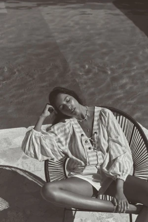 Monochrome photo of a woman in a striped shirt seated by a pool, eyes closed, bathed in sunlight, creating a relaxed and serene scene.