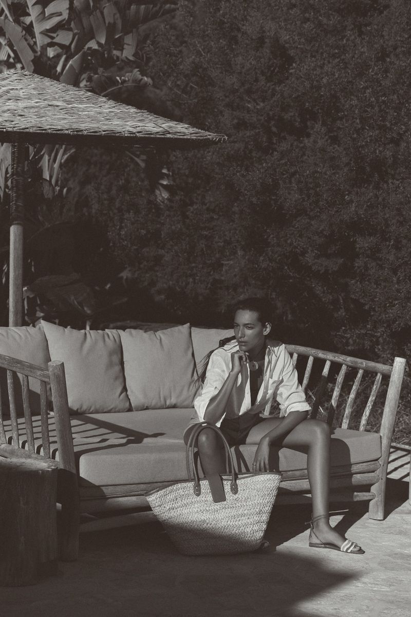 Monochrome portrait of a woman sitting on an outdoor couch under a shaded structure, featuring a woven bag and lush foliage backdrop.