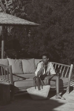 Monochrome portrait of a woman sitting on an outdoor couch under a shaded structure, featuring a woven bag and lush foliage backdrop.