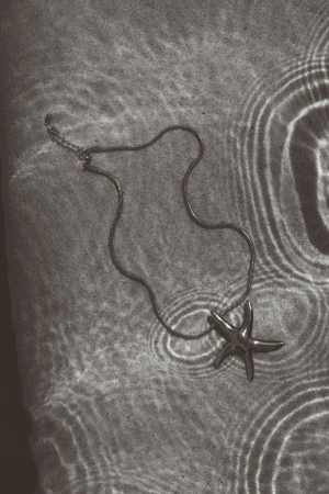 Starfish pendant on a silver necklace lays on a surface, with ripples of light creating a monochrome abstract pattern.