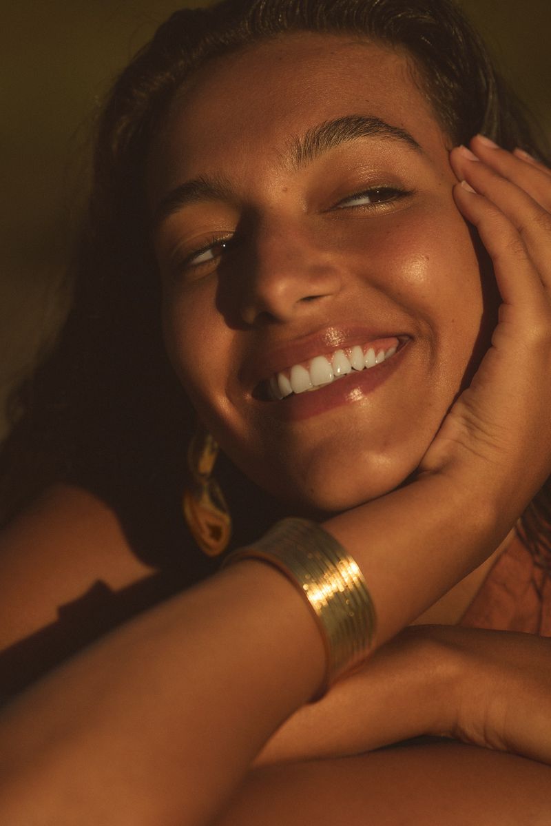 Close-up portrait of a smiling adult woman with golden skin, a gold cuff bracelet, and golden earrings, set against a blurred backdrop.