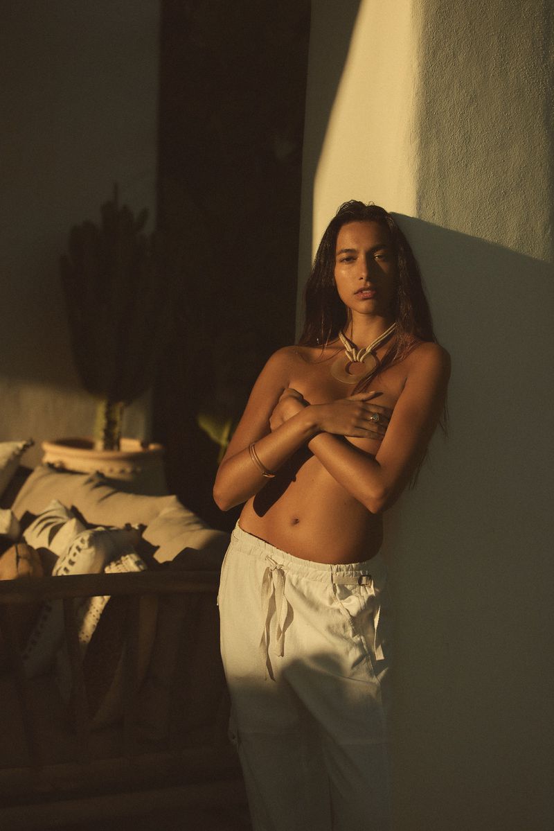 Young woman with long brown hair poses against a wall wearing white pants and minimal jewelry in natural, golden lighting.