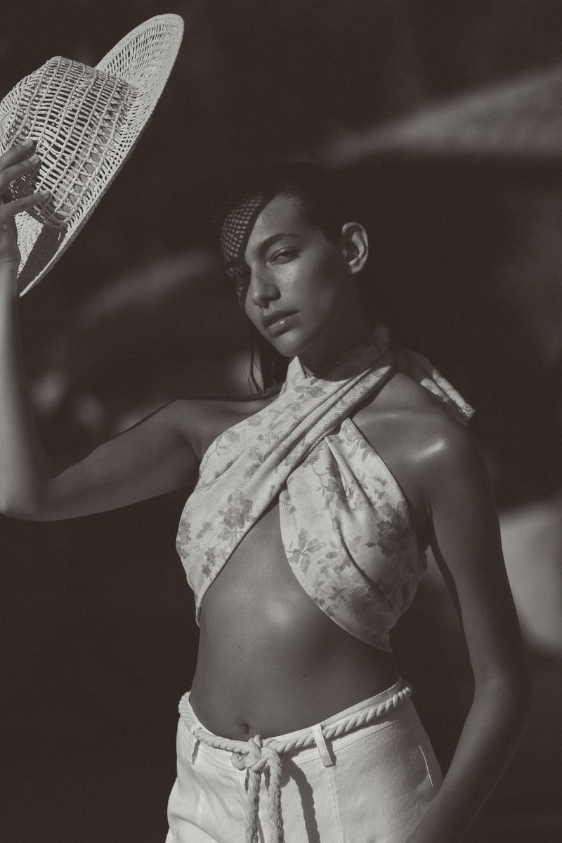 Monochrome portrait of a woman in a floral halter top and light jeans, holding a straw hat in a dark outdoor setting.