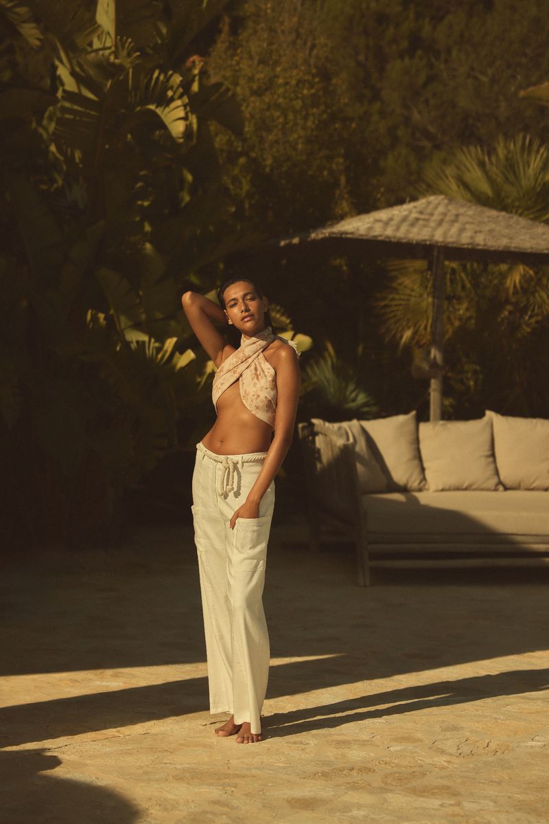 Woman poses in a tan floral wrap top and white linen pants in a tropical setting with lush green foliage and a lounge area.