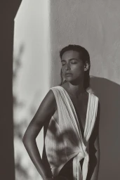 Monochrome portrait of a woman leaning against a stucco wall wearing a white sleeveless top with a knotted front.