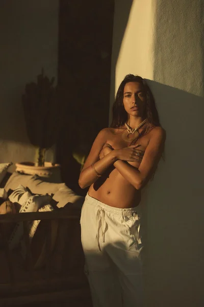 Model with long dark hair wearing white trousers and a statement necklace posing against a warm sunlit wall
