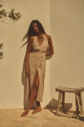 Model in a beige halter linen dress leaning against a white textured wall next to a wooden stool