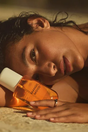 Close-up portrait of a woman reclining outdoors with golden hour sunlight hitting her face, holding a bottle of Mango Mood fragrance.