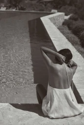 Woman in a white halter dress sitting by the edge of an outdoor swimming pool adjusting her straps