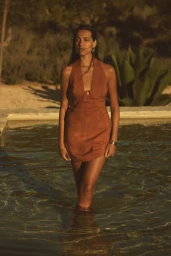Model wearing a rust halter dress standing waist-deep in an outdoor swimming pool during sunset.