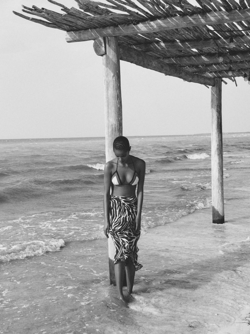 Black and white shot of an adult female standing in the water near a beach structure, wearing a bikini top and patterned sarong.
