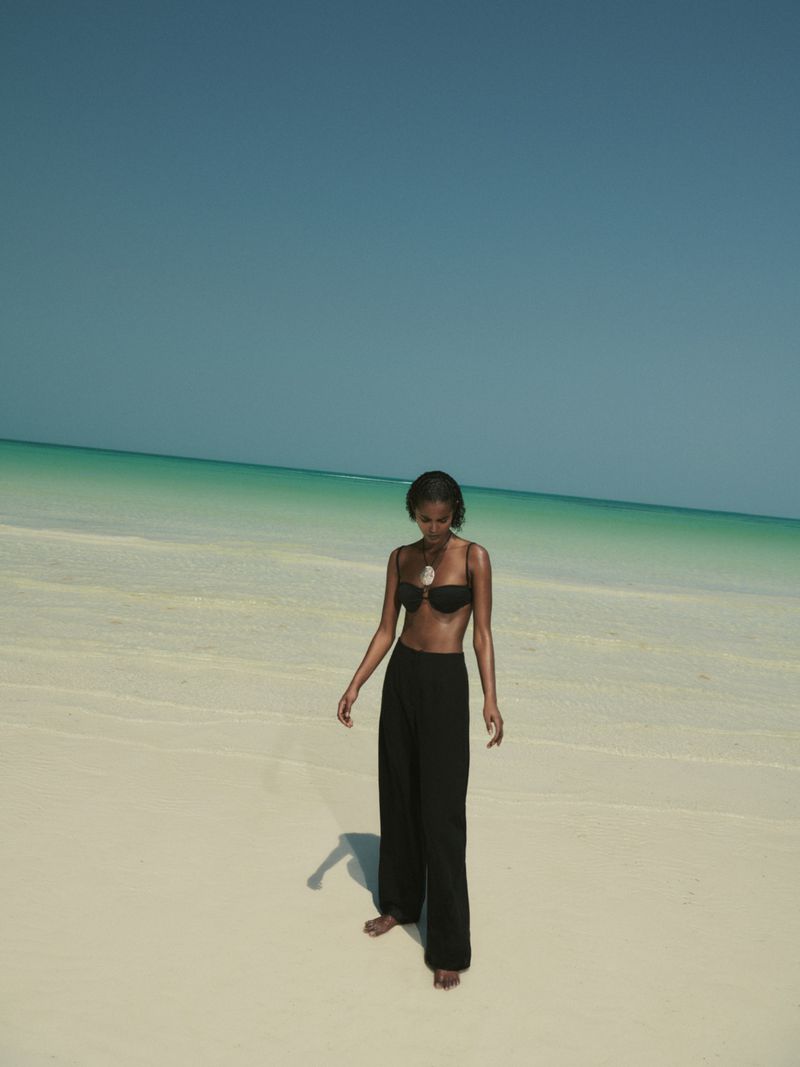 A dark-skinned woman stands barefoot on a sandy beach wearing a black bikini top and black wide-leg pants, under a blue sky.