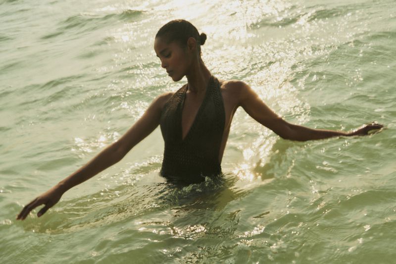 Adult female wading in ocean water, wearing dark halter swimsuit, wet hair in bun, arms extended, sunlit water background.