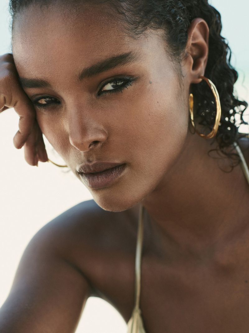 Close-up shot of a woman with dark hair and tan skin wearing hoop earrings. The model rests her head on her hand.