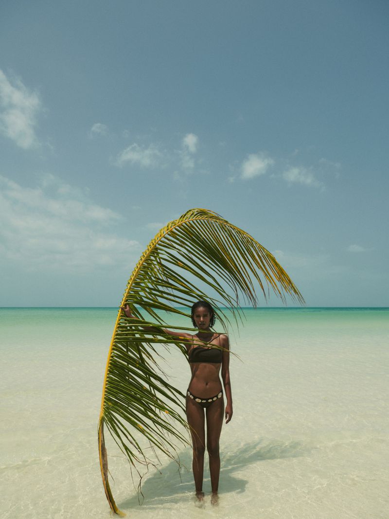 An adult woman stands in shallow sea water on a sunny day, holding a large palm frond to her right, with the ocean and sky behind her.