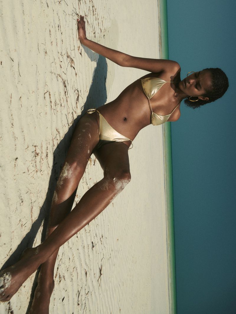 Woman in a gold bikini poses on a sandy beach with a bright blue sky in the background. Outdoor fashion and summer vibes.
