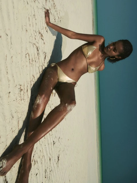 Woman in a gold bikini poses on a sandy beach with a bright blue sky in the background. Outdoor fashion and summer vibes.
