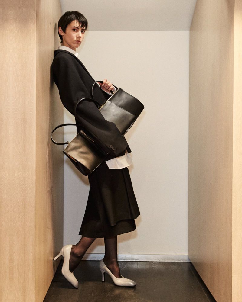 Elegant woman in a black blazer and skirt holding black handbags, leaning against a wall in stylish high-heeled shoes.