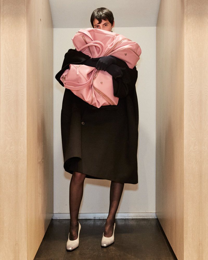 A fashionable person poses indoors with three large pink tote bags, wearing a dark coat, black tights, and cream-colored pumps.