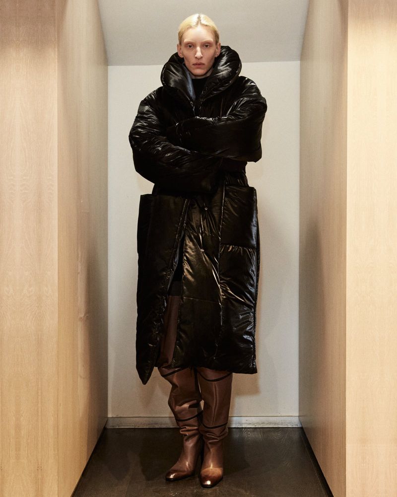 Full-length fashion portrait of a slim adult woman wearing a shiny black puffer coat and shiny brown boots in an enclosed hallway.
