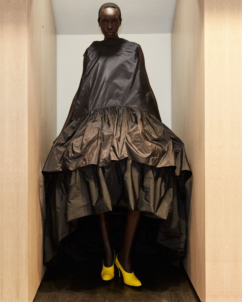 A dark-skinned woman poses in a dramatic black dress and yellow heels against a neutral interior, highlighting fashion and style.