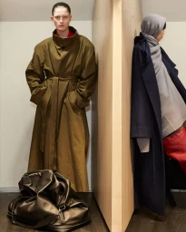 Two models showcasing oversized coats by Marie Adam-Leenaerdt, with a large dark duffel bag on the floor in an interior setting.