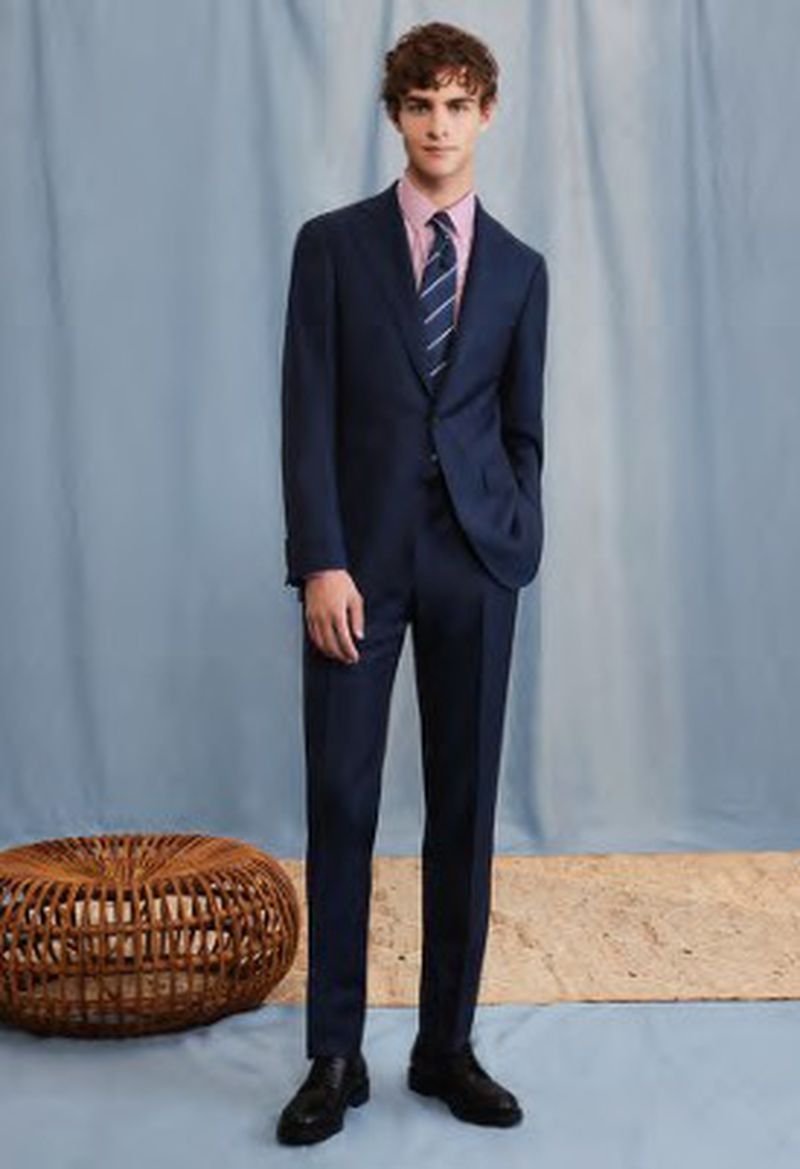 Man in a navy suit, pink shirt, and striped tie standing in a studio setting with blue background, tan flooring, and woven ottoman.