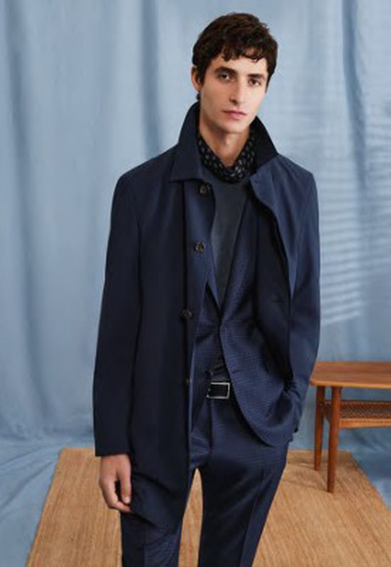 Fashion shot of a man in a suit and overcoat, posing indoors against a draped backdrop with a mid-century modern side table.