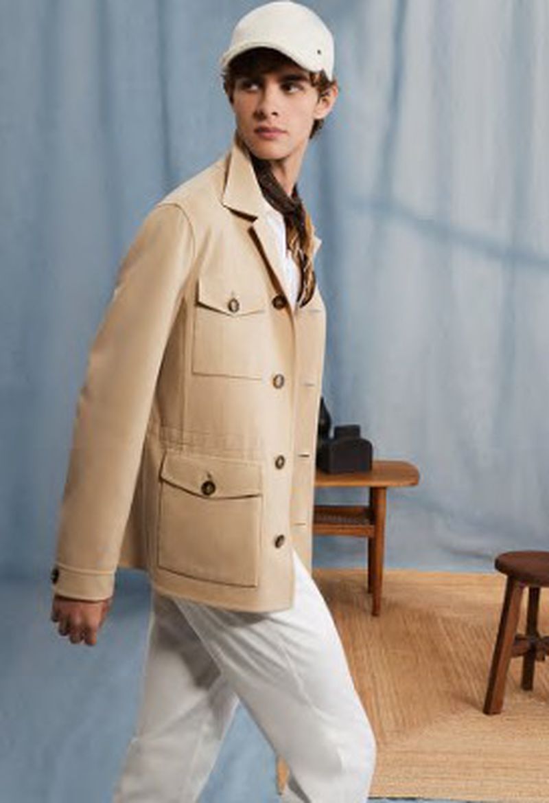 Man in safari jacket and cap poses indoors against a light blue backdrop with tables, white pants, tan jacket, and tan cap.