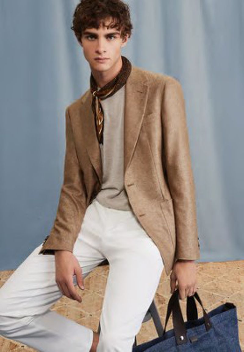 A stylish adult male wearing a light brown jacket, light-colored pants, and a patterned scarf poses with a blue tote bag.