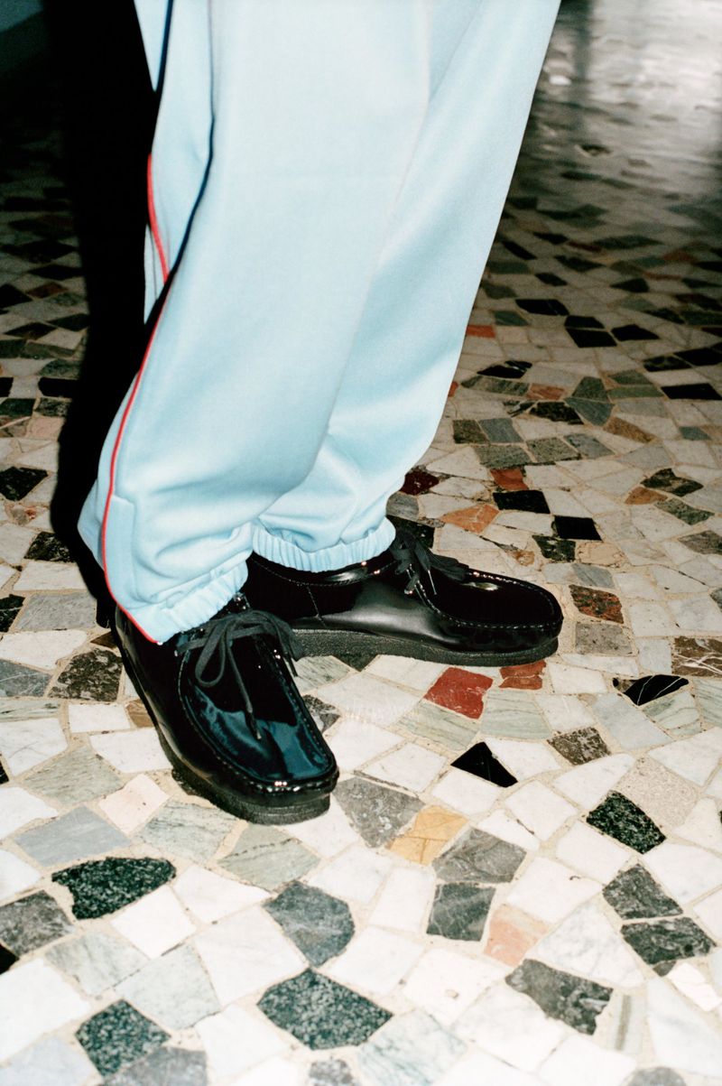 A person wearing baby-blue tracksuit pants and black patent leather shoes stands on a tessellated marble floor in an indoor setting.