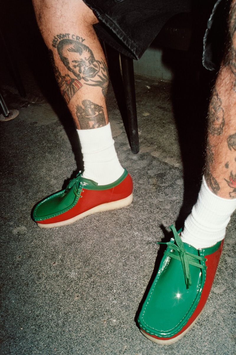 Person wearing red and green leather shoes and white socks, showing tattoos on lower legs and feet on gray concrete.