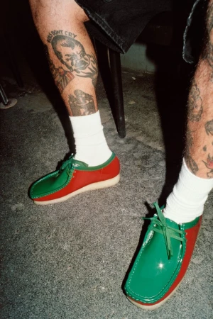 Person wearing red and green leather shoes and white socks, showing tattoos on lower legs and feet on gray concrete.