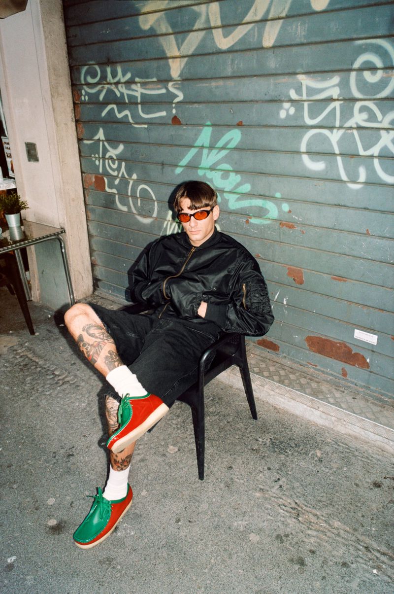 Man in black jacket and shorts sits on a black chair outdoors against graffiti-covered wall, wearing red sunglasses and colorful shoes.