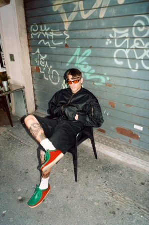Man in black jacket and shorts sits on a black chair outdoors against graffiti-covered wall, wearing red sunglasses and colorful shoes.