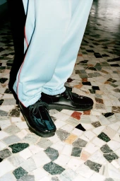 Close-up of black leather lace-up shoes and light blue trousers standing on a patterned mosaic floor.