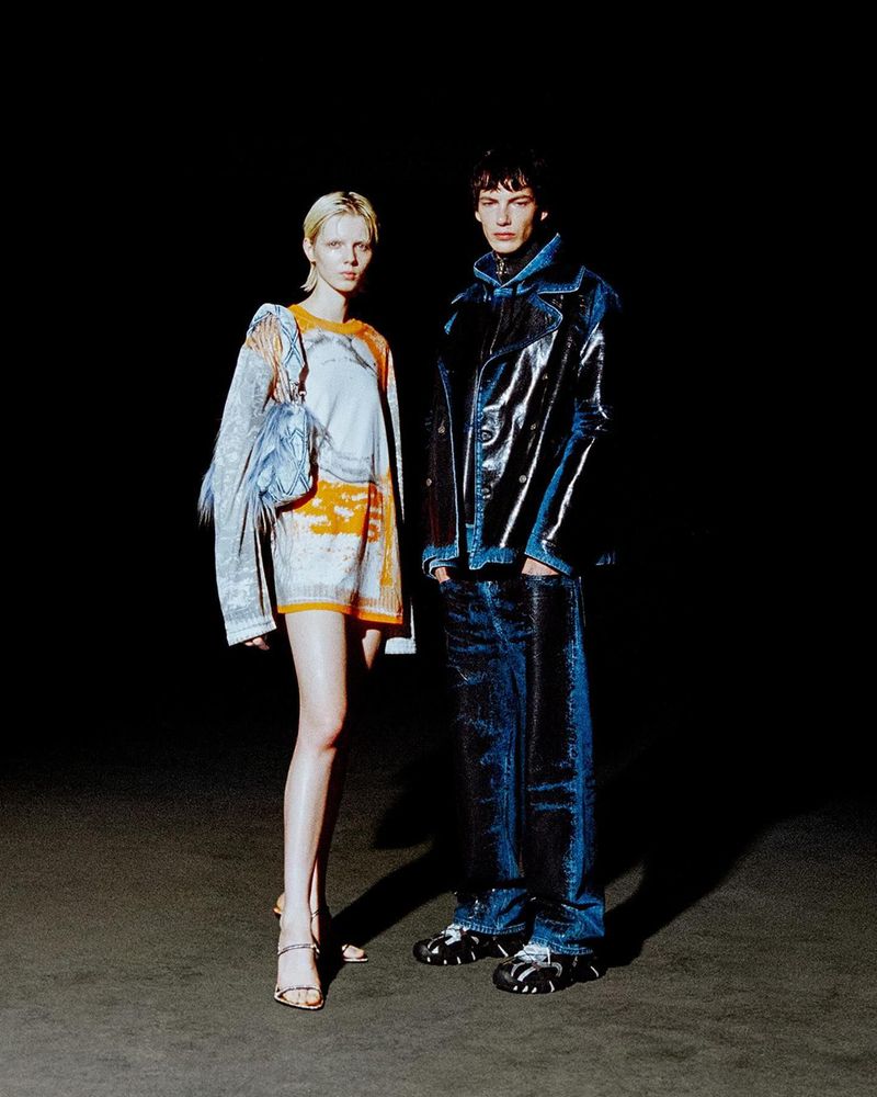 Two fashion models, one female and one male, stand in a dark studio. The female wears a dress and the male wears a jacket and jeans.