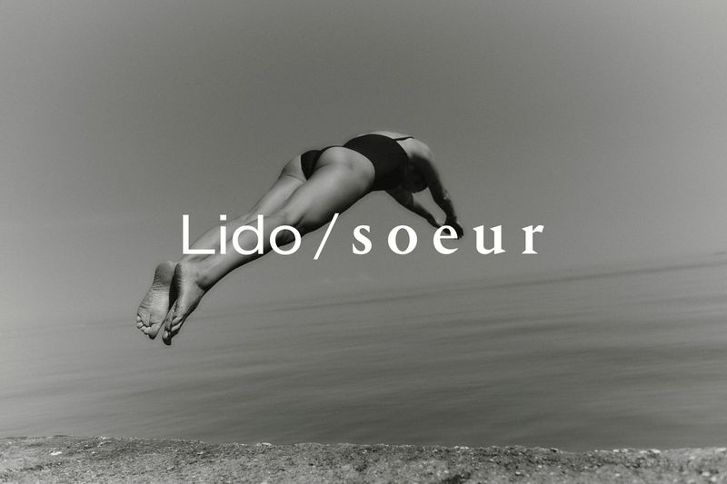Monochrome image of a woman in a swimsuit diving into the ocean, captured with a low angle against a clear sky and horizon line.
