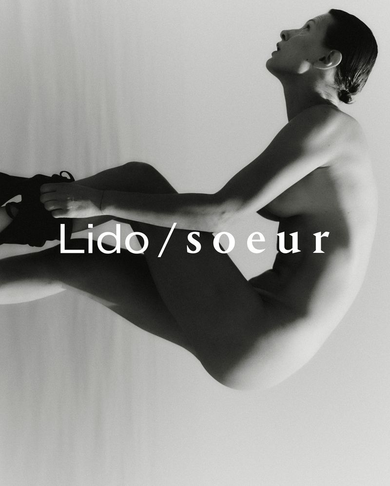 Black and white fashion photograph of a nude woman sitting in an artistic pose against a white background with dark shoes on.
