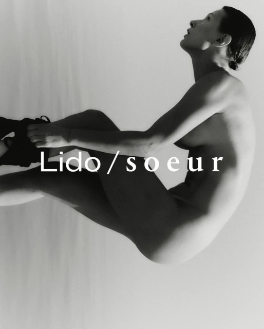 Black and white fashion photograph of a nude woman sitting in an artistic pose against a white background with dark shoes on.