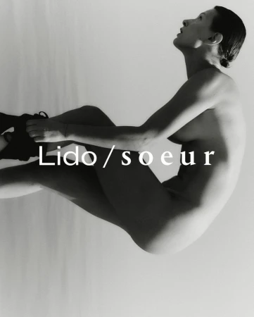 Black and white fashion photograph of a nude woman sitting in an artistic pose against a white background with dark shoes on.