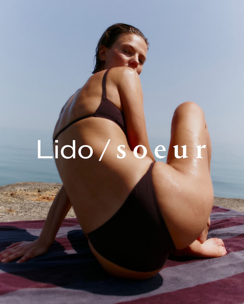 Woman poses on beach towel wearing a brown bikini. Clear blue sky and water in background. Wet skin and minimal makeup.